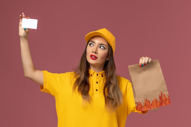 Vista frontal del mensajero femenino en uniforme amarillo y gorra con paquete de alimentos y tarjeta de plástico en la pared rosa