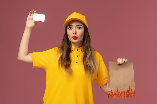 Vista frontal del mensajero femenino en uniforme amarillo y gorra con paquete de alimentos y tarjeta de plástico en la pared rosa