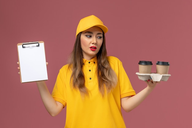 Vista frontal del mensajero femenino en uniforme amarillo y gorra sosteniendo el bloc de notas de tazas de café de entrega en la pared rosa claro