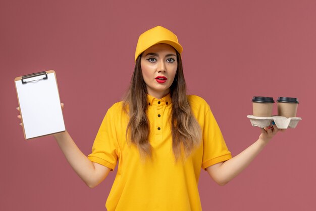 Vista frontal del mensajero femenino en uniforme amarillo y gorra sosteniendo el bloc de notas de tazas de café de entrega en la pared rosa claro