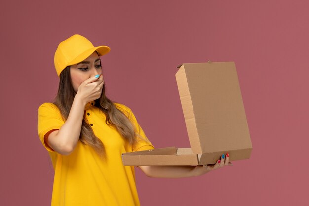 Vista frontal del mensajero femenino en uniforme amarillo y gorra sosteniendo una caja de comida abierta con expresión de sorpresa en la pared rosa