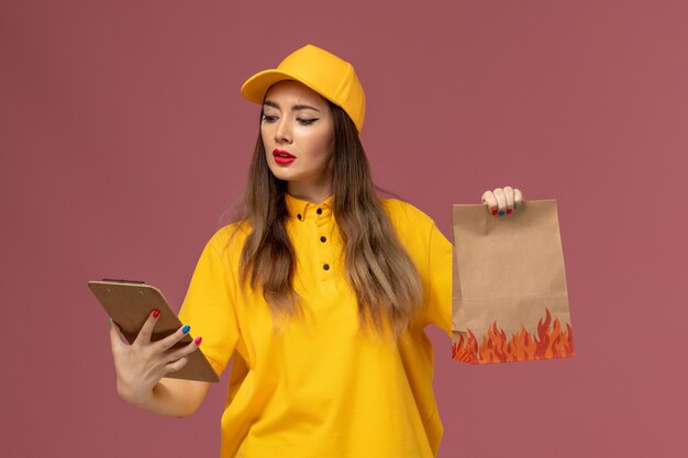 Vista frontal del mensajero femenino en uniforme amarillo y gorra sosteniendo el paquete de alimentos y el bloc de notas en la pared rosa