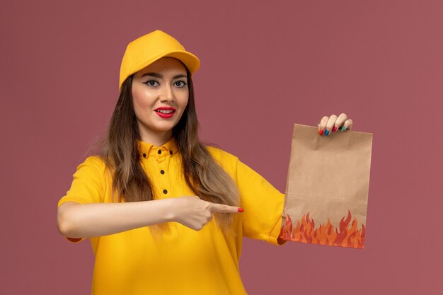 Vista frontal del mensajero femenino en uniforme amarillo y gorra sosteniendo el paquete de alimentos en la pared rosa claro