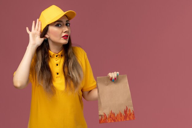 Vista frontal del mensajero femenino en uniforme amarillo y gorra sosteniendo el paquete de alimentos tratando de escuchar en la pared de color rosa claro