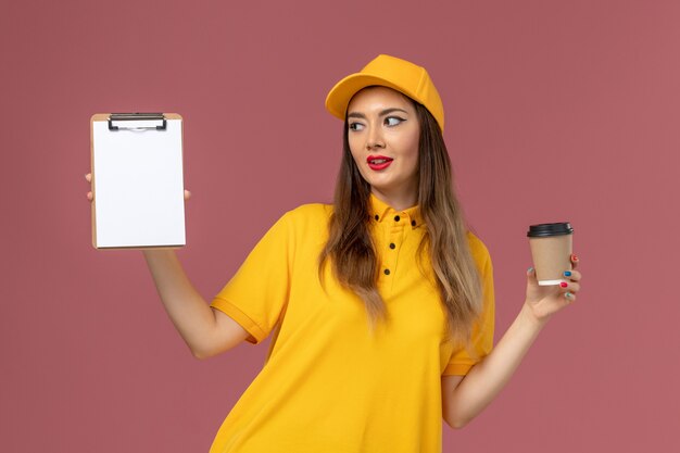 Vista frontal del mensajero femenino en uniforme amarillo y gorra sosteniendo la taza de café de entrega y el bloc de notas en la pared rosa