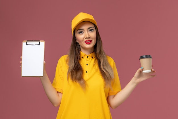 Vista frontal del mensajero femenino en uniforme amarillo y gorra sosteniendo la taza de café de entrega y el bloc de notas en la pared rosa