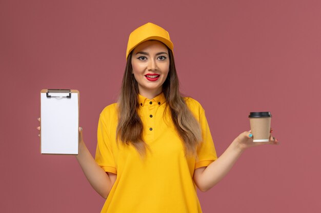 Vista frontal del mensajero femenino en uniforme amarillo y gorra sosteniendo la taza de café de entrega y el bloc de notas en la pared rosa