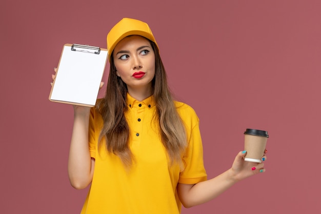 Vista frontal del mensajero femenino en uniforme amarillo y gorra sosteniendo la taza de café de entrega y el bloc de notas pensando en la pared rosa