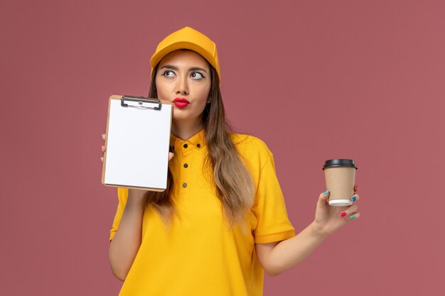 Vista frontal del mensajero femenino en uniforme amarillo y gorra sosteniendo la taza de café de entrega y el bloc de notas pensando en la pared rosa