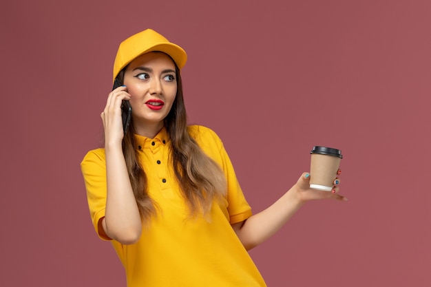 Vista frontal del mensajero femenino en uniforme amarillo y gorra sosteniendo la taza de café de entrega y hablando por teléfono en la pared rosa