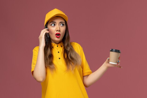 Vista frontal del mensajero femenino en uniforme amarillo y gorra sosteniendo la taza de café de entrega y hablando por teléfono en la pared rosa