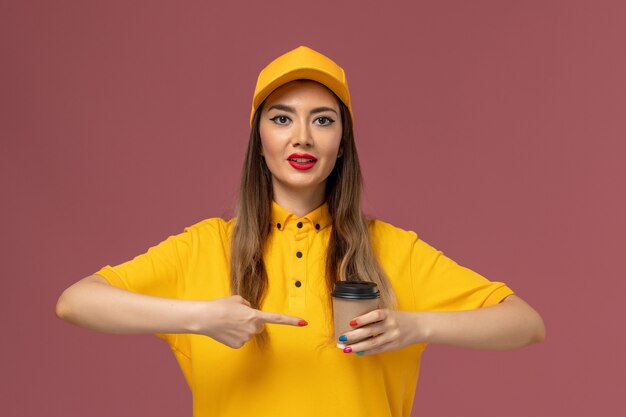 Vista frontal del mensajero femenino en uniforme amarillo y gorra sosteniendo la taza de café de entrega en la pared rosa