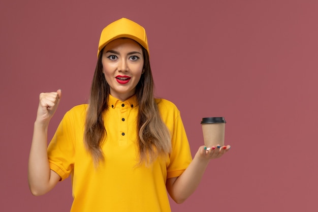 Vista frontal del mensajero femenino en uniforme amarillo y gorra sosteniendo la taza de café de entrega en la pared rosa