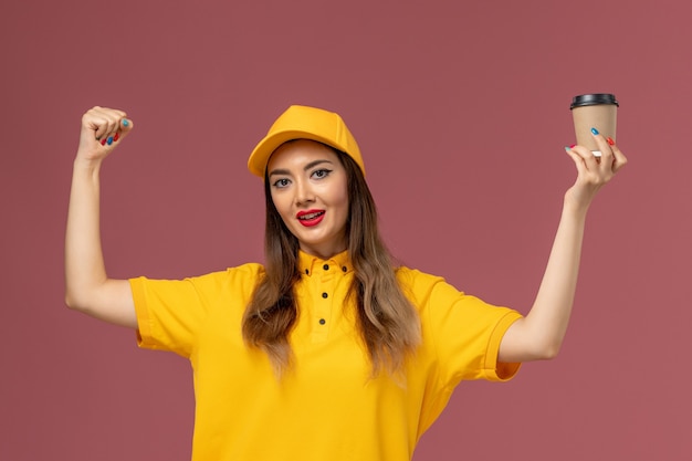 Vista frontal del mensajero femenino en uniforme amarillo y gorra sosteniendo la taza de café de entrega y regocijándose en la pared rosa