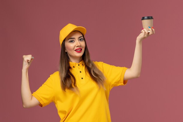 Vista frontal del mensajero femenino en uniforme amarillo y gorra sosteniendo la taza de café de entrega con una sonrisa en la pared rosa