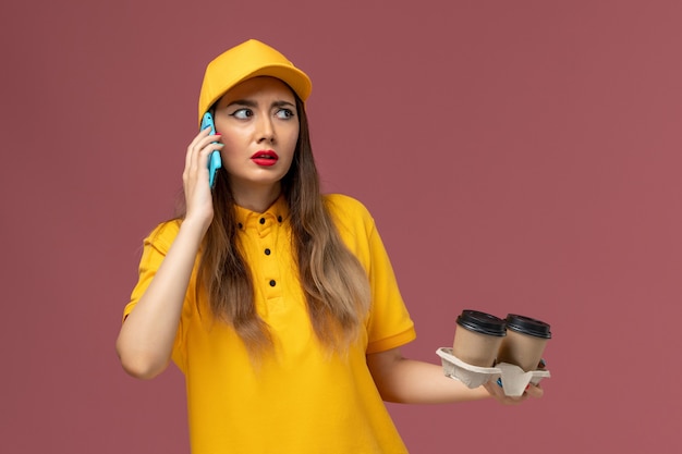 Vista frontal del mensajero femenino en uniforme amarillo y gorra sosteniendo tazas de café de entrega hablando por teléfono en la pared rosa