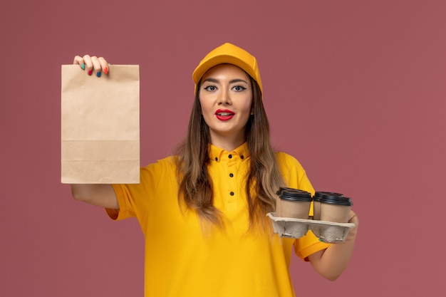 Vista frontal del mensajero femenino en uniforme amarillo y gorra sosteniendo tazas de café de entrega marrón y paquete de comida en la pared rosa