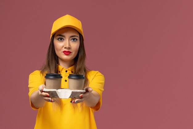 Vista frontal del mensajero femenino en uniforme amarillo y gorra sosteniendo tazas de café de entrega marrón en la pared rosa