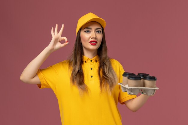 Vista frontal del mensajero femenino en uniforme amarillo y gorra sosteniendo tazas de café de entrega marrón en la pared rosa