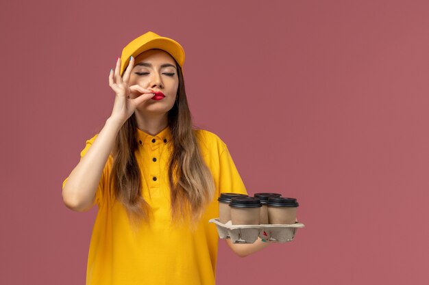 Vista frontal del mensajero femenino en uniforme amarillo y gorra sosteniendo tazas de café de entrega marrón en la pared rosa