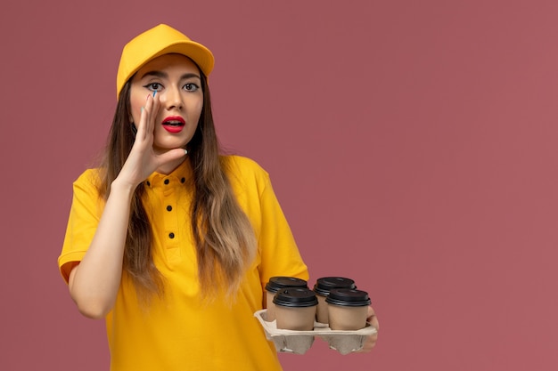 Vista frontal del mensajero femenino en uniforme amarillo y gorra sosteniendo tazas de café de entrega marrón susurrando en la pared rosa