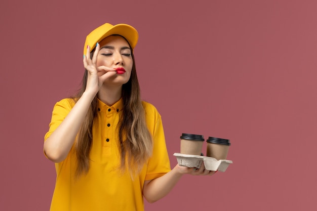 Vista frontal del mensajero femenino en uniforme amarillo y gorra sosteniendo tazas de café de entrega en la pared rosa