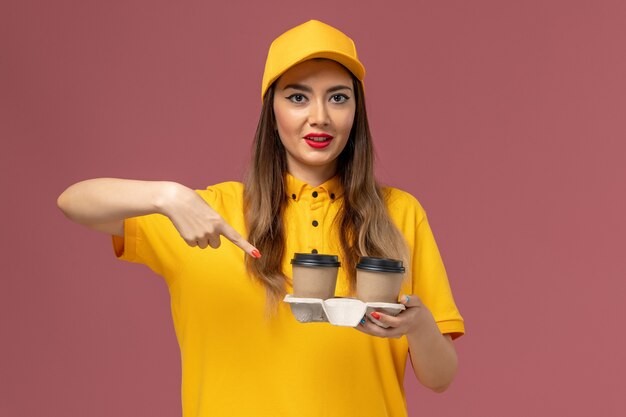 Vista frontal del mensajero femenino en uniforme amarillo y gorra sosteniendo tazas de café de entrega en la pared rosa