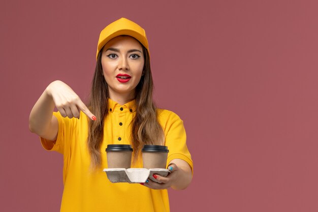 Vista frontal del mensajero femenino en uniforme amarillo y gorra sosteniendo tazas de café de entrega en la pared rosa