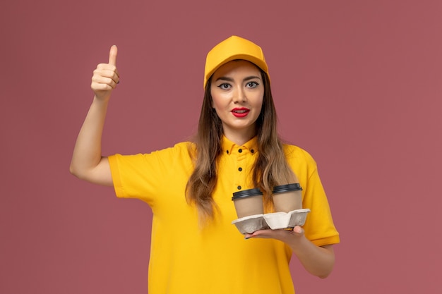 Vista frontal del mensajero femenino en uniforme amarillo y gorra sosteniendo tazas de café de entrega en la pared rosa