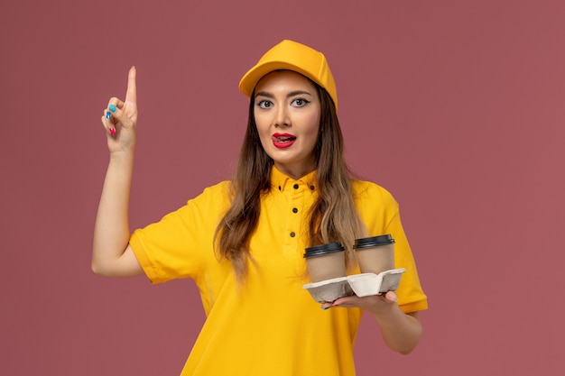 Vista frontal del mensajero femenino en uniforme amarillo y gorra sosteniendo tazas de café de entrega en la pared rosa