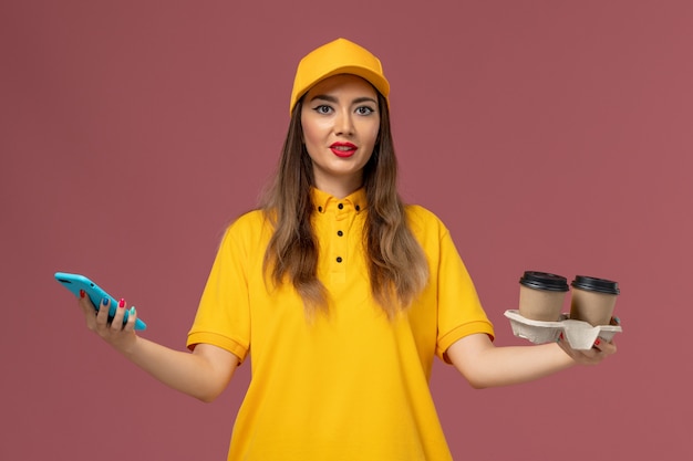 Vista frontal del mensajero femenino en uniforme amarillo y gorra sosteniendo tazas de café de entrega y teléfono inteligente en la pared rosa