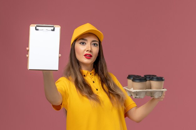 Vista frontal del mensajero femenino en uniforme amarillo y gorra sosteniendo tazas de café marrón y bloc de notas en la pared rosa