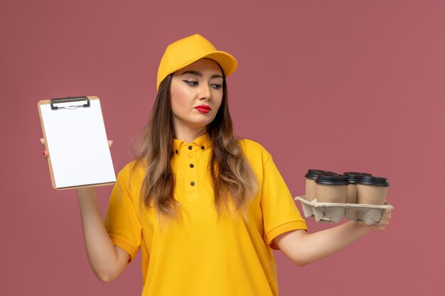 Vista frontal del mensajero femenino en uniforme amarillo y gorra sosteniendo tazas de café marrón y bloc de notas en la pared rosa