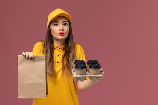 Vista frontal del mensajero femenino en uniforme amarillo y gorra sosteniendo tazas de café marrón y paquete de comida en la pared rosa