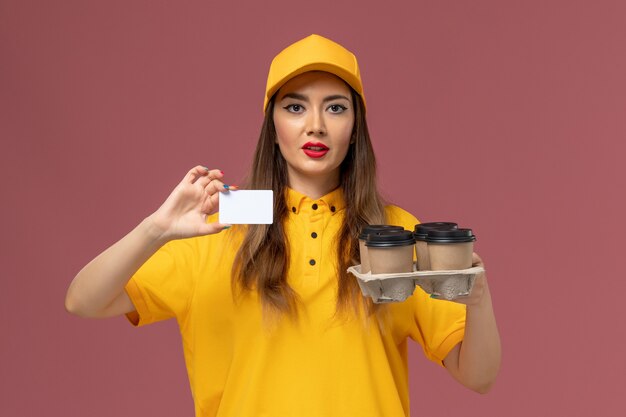 Vista frontal del mensajero femenino en uniforme amarillo y gorra sosteniendo tazas de café marrón y tarjeta en la pared rosa