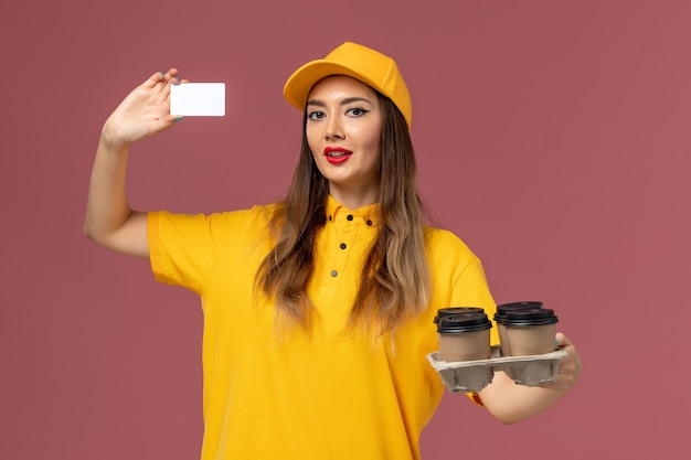 Vista frontal del mensajero femenino en uniforme amarillo y gorra sosteniendo tazas de café marrón y tarjeta en la pared rosa