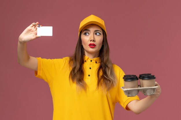 Vista frontal del mensajero femenino en uniforme amarillo y gorra sosteniendo tazas de café marrón y tarjeta en la pared rosa