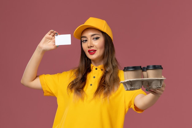 Vista frontal del mensajero femenino en uniforme amarillo y gorra sosteniendo tazas de café marrón y tarjeta en la pared rosa