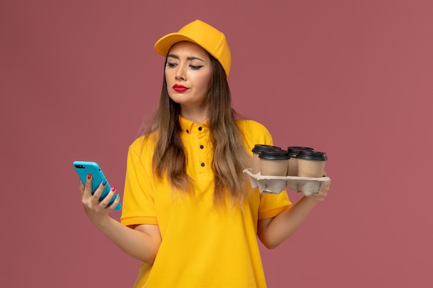 Foto gratuita vista frontal del mensajero femenino en uniforme amarillo y gorra sosteniendo tazas de café marrón y usando su teléfono en la pared rosa