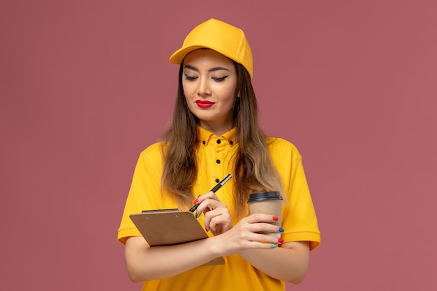Vista frontal del mensajero femenino en uniforme amarillo y gorra con taza de café de entrega y bloc de notas con bolígrafo en la pared rosa
