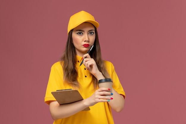 Vista frontal del mensajero femenino en uniforme amarillo y gorra con taza de café de entrega y bloc de notas con bolígrafo en la pared rosa