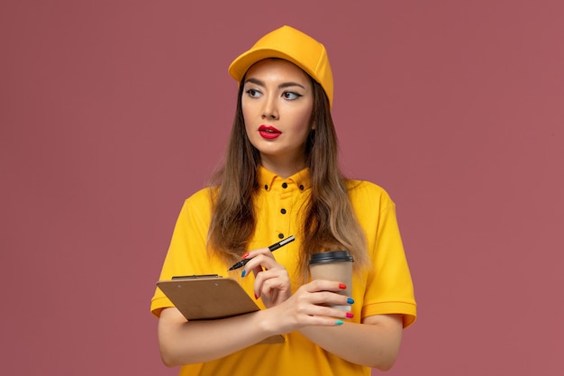 Vista frontal del mensajero femenino en uniforme amarillo y gorra con taza de café de entrega y bloc de notas con bolígrafo