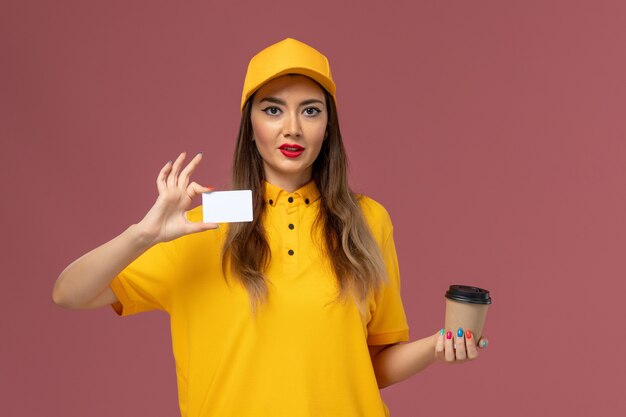 Vista frontal del mensajero femenino en uniforme amarillo y gorra con taza de café de entrega y tarjeta blanca en la pared rosa