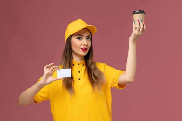 Vista frontal del mensajero femenino en uniforme amarillo y gorra con taza de café de entrega y tarjeta blanca en la pared rosa