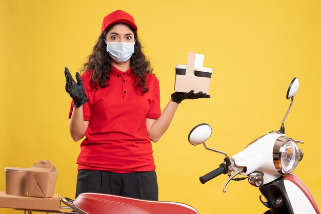 Vista frontal mensajero femenino en uniforme rojo sosteniendo tazas de café sobre fondo amarillo trabajador entrega covid- uniforme de trabajo pandémico