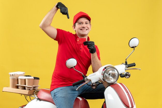 Vista frontal del mensajero hombre vestido con blusa roja y guantes de sombrero en máscara médica entregando orden sentado en scooter apuntando con tarjeta bancaria