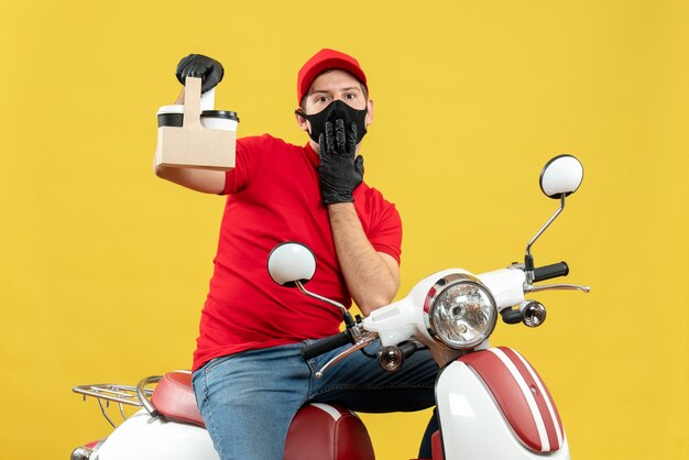 Vista frontal del mensajero hombre vestido con blusa roja y sombrero guantes en máscara médica sentado en scooter sosteniendo órdenes sintiéndose sorprendido