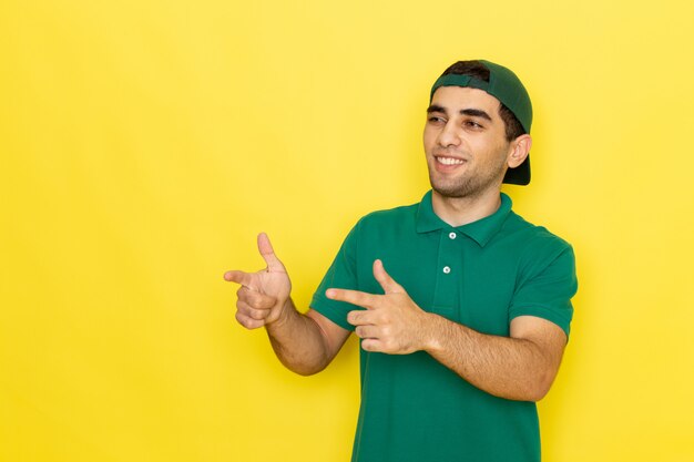 Vista frontal, mensajero, joven, macho, en, camisa verde, gorra verde, sonriente, y, señalar, el suyo, dedos, en, el, fondo amarillo, trabajo, entregar, servicio, color