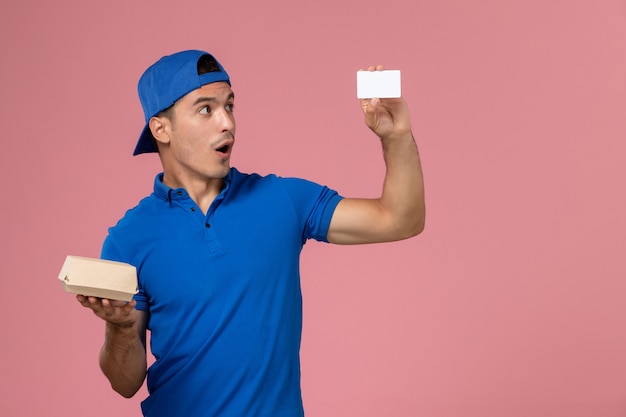 Vista frontal, mensajero, macho joven, en, uniforme azul, capa, tenencia, pequeño, entrega, paquete de comida, y, blanco, tarjeta, en, pared rosa claro