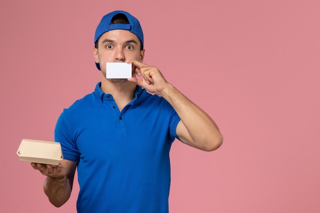 Vista frontal, mensajero, macho joven, en, uniforme azul, capa, tenencia, pequeño, entrega, paquete de comida, y, blanco, tarjeta, en, pared rosa claro